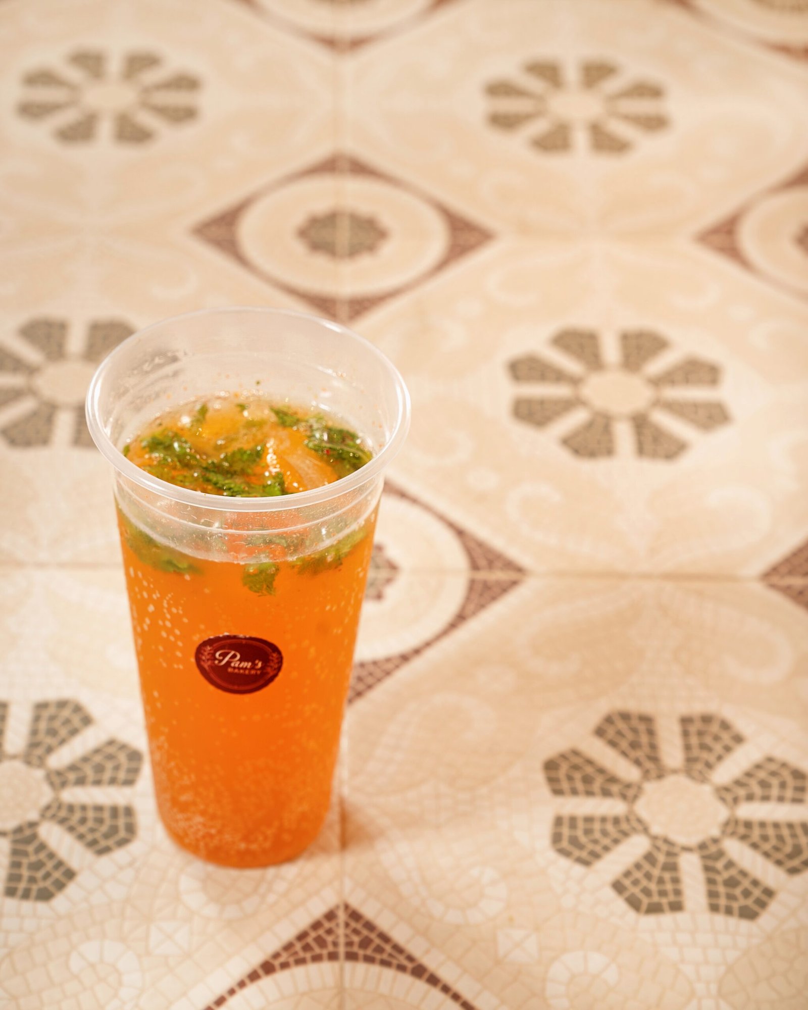 Orange fizzy beverage with fresh mint and citrus garnish served in a branded cup at Pam’s Bakery.