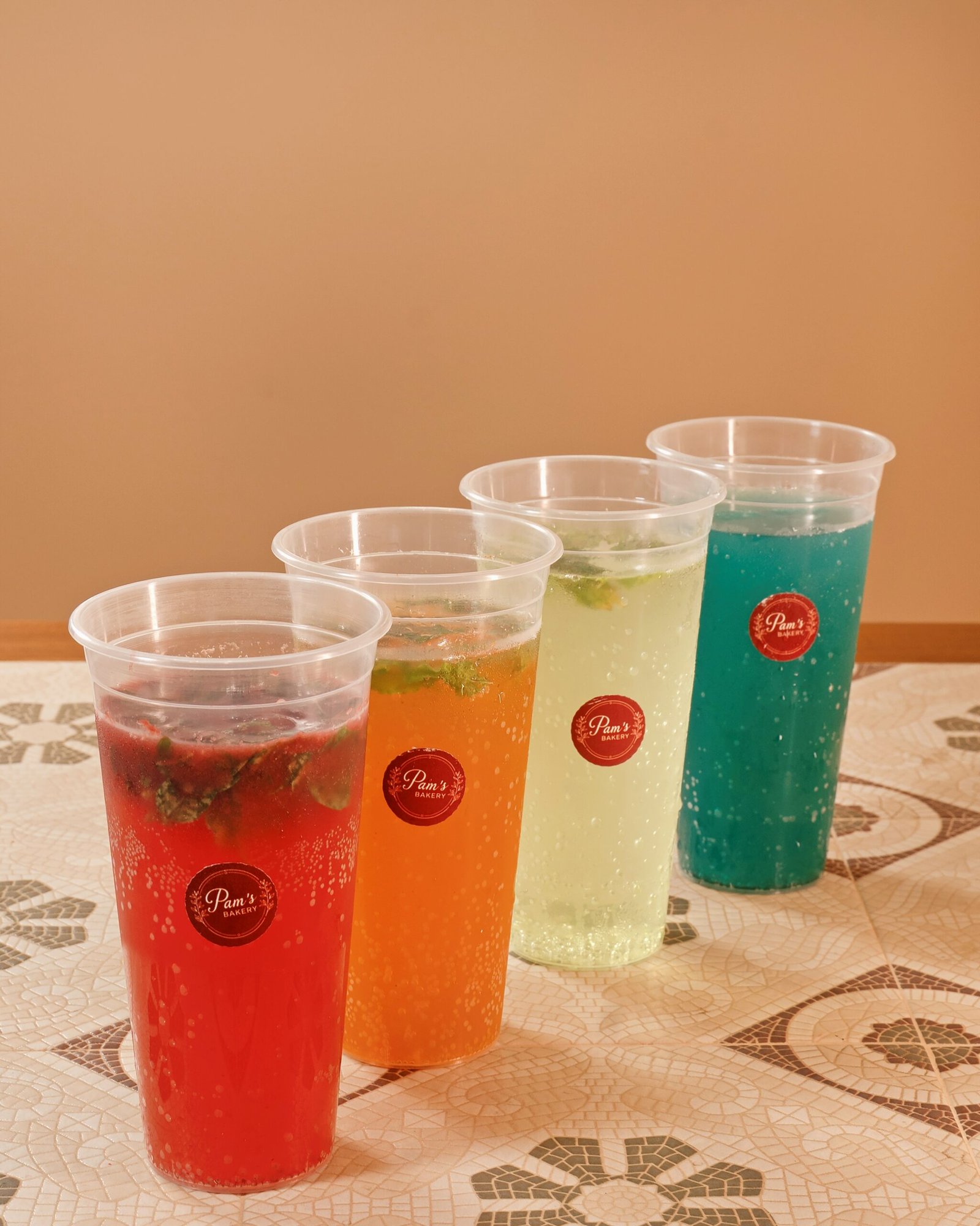 Four colorful beverages served in branded cups at Pam’s Bakery, arranged from red and orange to pale green and blue.