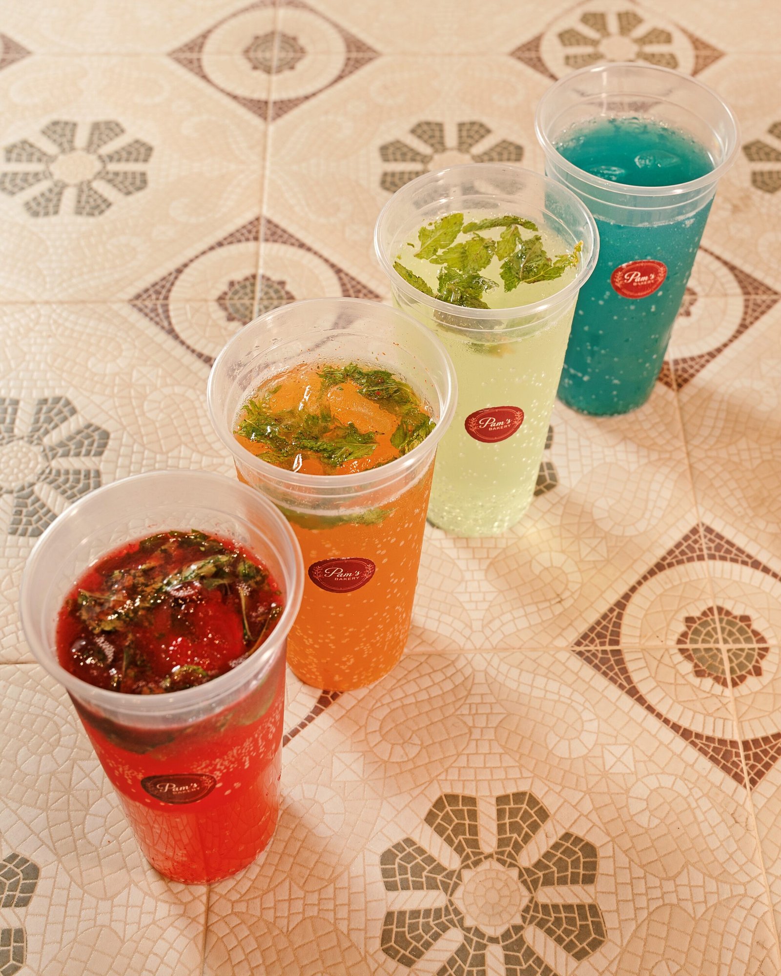 Four colorful beverages served in branded cups at Pam’s Bakery, including red, orange, green, and blue drinks.